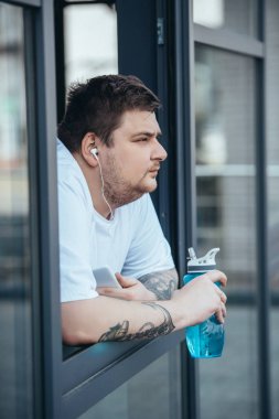 kulaklık ve akıllı telefon spor şişesi tutan ve spor salonunda pencereden dışarı bakarak kilolu adam