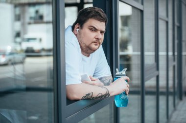 kulaklık ve akıllı telefon spor şişesi tutan ve spor salonunda pencereden dışarı bakarak obez adam
