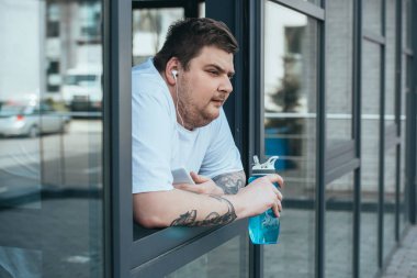 kulaklık ve akıllı telefon spor şişesi tutan ve spor salonunda pencereden dışarı bakarak kilolu adam