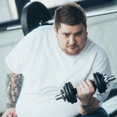 Kilolu dövmeli adam Kamera bakıyor ve spor merkezinde dumbbell ile egzersiz
