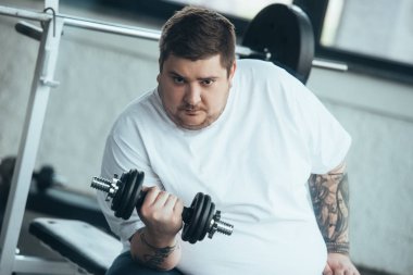 Kilolu dövmeli adam Kamera bakıyor ve spor merkezinde dumbbell ile eğitim