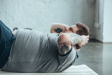 kilolu dövmeli adam fitness mat üzerinde yatan ve spor merkezinde egzersiz kadar oturup yapıyor