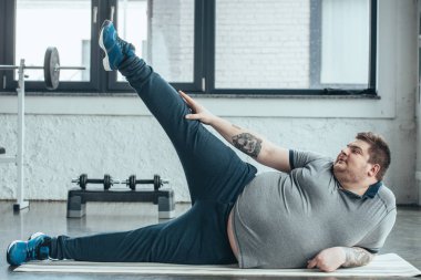 fitness mat ve spor salonunda bacaklar germe yatan kilolu dövmeli adam