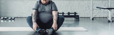 fitness mat üzerinde oturan kilolu adam panoramik çekim ve spor merkezinde bacaklar germe