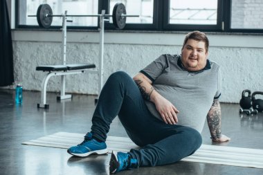 aşırı kilolu dövmeli adam kamera bakıyor ve spor merkezinde fitness mat oturuyor