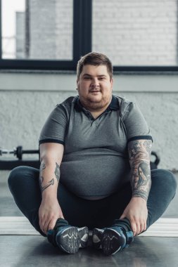 overweight tattooed man sitting and Looking At Camera during stretching exercise at gym clipart