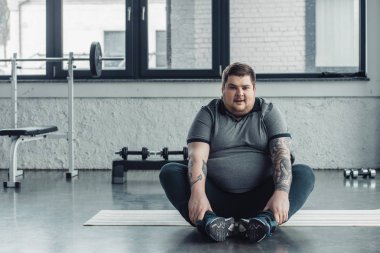 kilolu dövmeli adam oturma ve spor salonunda germe egzersiz sırasında Kamera Bakıyor