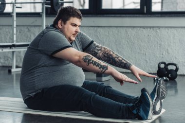 kilolu dövmeli adam fitness mat üzerinde oturan ve spor salonunda germe