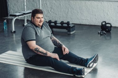 spor merkezinde fitness mat üzerinde oturan gri t-shirt kilolu dövmeli adam