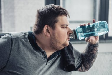 overweight tattooed man drinking water from sport bottle at sports center clipart