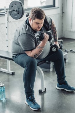 Kilolu dövmeli adam oturma ve spor merkezinde dumbbells ile egzersiz