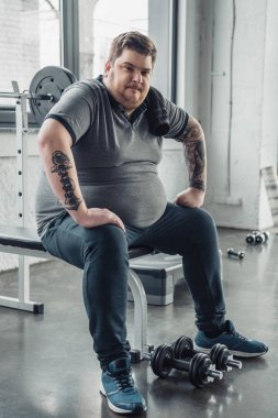 yorgun Obez adam bankta oturan ve spor salonunda halter ile egzersiz yaptıktan sonra Kamera Bakıyor