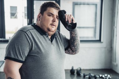tired overweight tattooed man looking at camera and wiping face with towel at sports center clipart