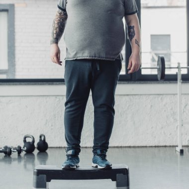 spor merkezinde adım platformu üzerinde duran kilolu dövmeli adam kırpılmış görünümü