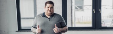 panoramic shot of overweight tattooed man Looking At Camera and showing thumbs up  clipart