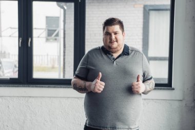 overweight tattooed man in grey t-shirt Looking At Camera and showing thumbs up clipart