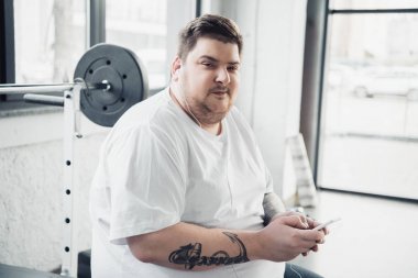 kulaklıklı kilolu adam bankta oturan, kamera bakarak ve spor salonunda akıllı telefon kullanarak
