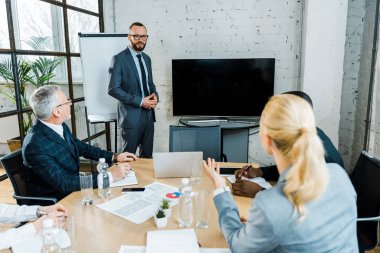  business coach in formal wear standing near tv with blank screen and multicultural coworkers 