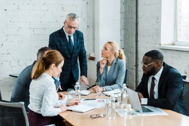 business coach in formal wear standing near multicultural coworkers on meeting clipart