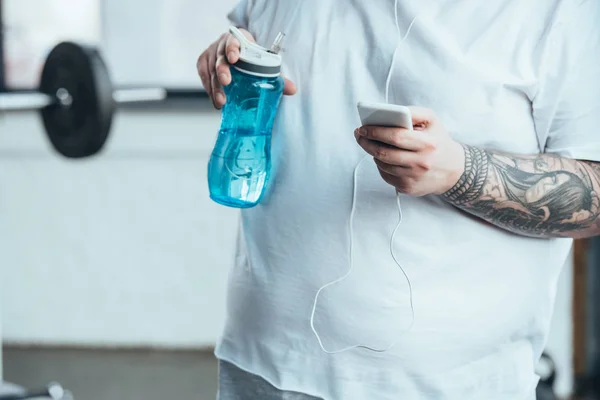 裁剪视图超重纹身男子拿着运动瓶和使用智能手机在健身房 — 图库照片