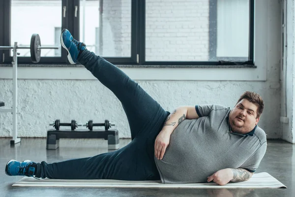 超重纹身男子躺在健身垫和伸展腿在健身房 — 图库照片