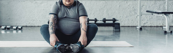 panoramic shot of overweight man sitting on fitness mat and stretching legs at sports center