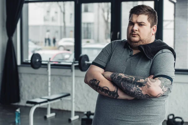 overweight tattooed man with towel and crossed arms looking at camera at gym