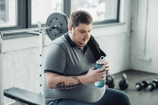 超重纹身男子坐在长凳上 拿着运动瓶在健身房 — 图库照片