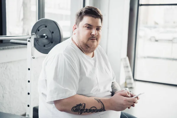 overweight man in earphones sitting on bench, looking at camera and using smartphone at gym