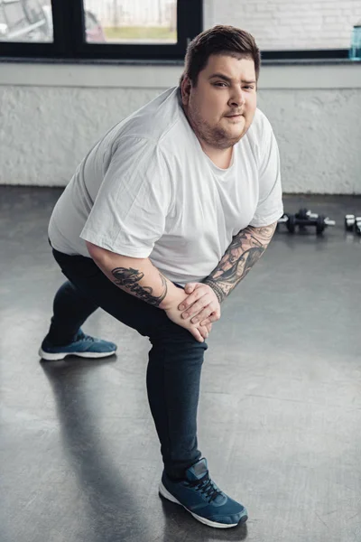 overweight tattooed man looking at camera while stretching legs at sports center