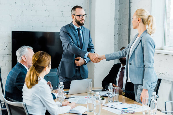 handsome businessman and  blonde businesswoman shaking hands near multicultural coworkers 