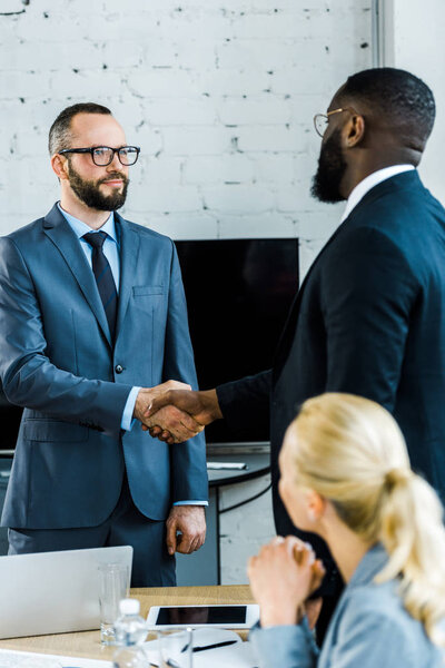 multicultural businessmen shaking hands near blonde coworker in office 