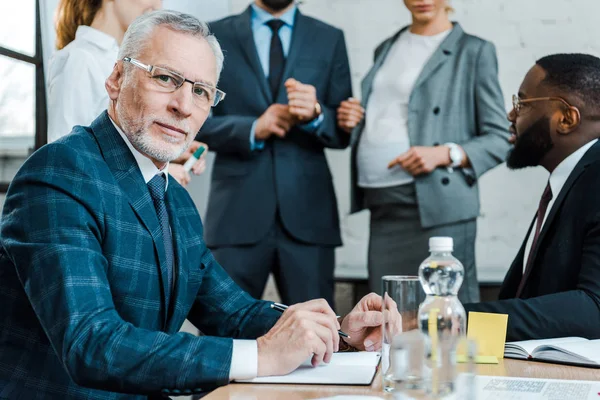 Selektiv Fokus Skäggiga Affärsman Glasögon Tittar Kamera Nära Mångkulturella Medarbetare — Stockfoto