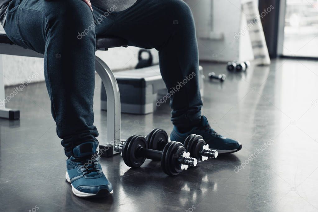 cropped view of man in trainers sitting with dumbbells at sports center