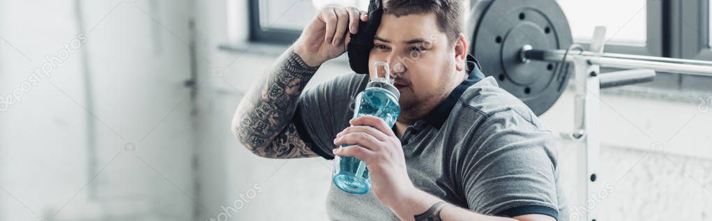 panoramic shot of overweight tattooed man drinking water from sport bottle and wiping face with towel at gym