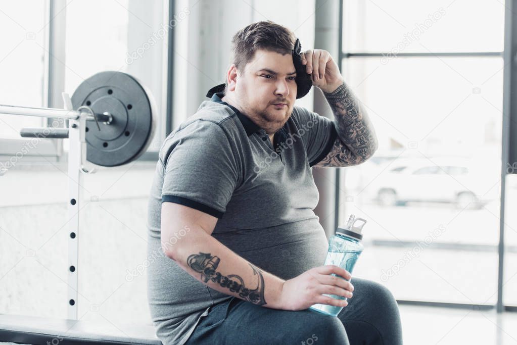overweight tattooed man holding sport bottle and wiping face with towel at gym