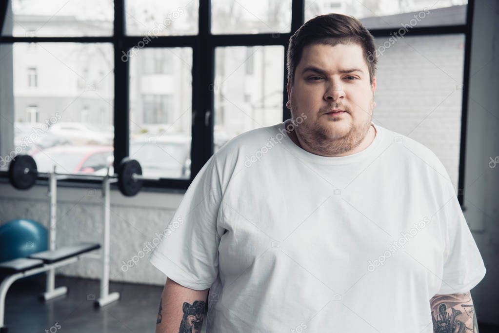overweight tattooed man in white t-shirt looking at camera at sports center with copy space