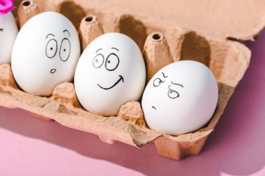 eggs with surprised, angry and smiling face expressions in egg carton on pink clipart