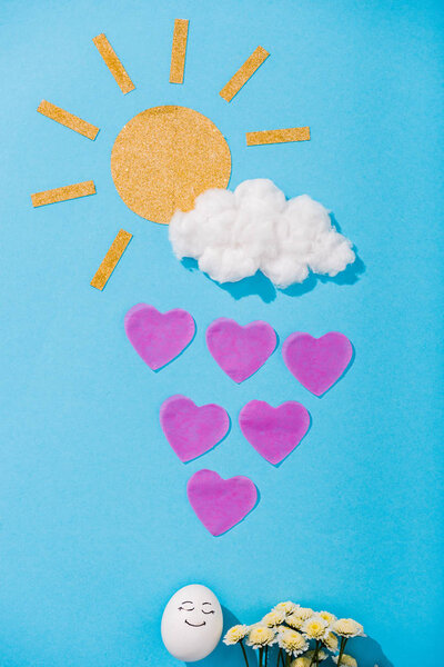 top view of paper sun, cotton candy cloud, egg with happy face expression, flowers and heart-shaped raindrops on blue