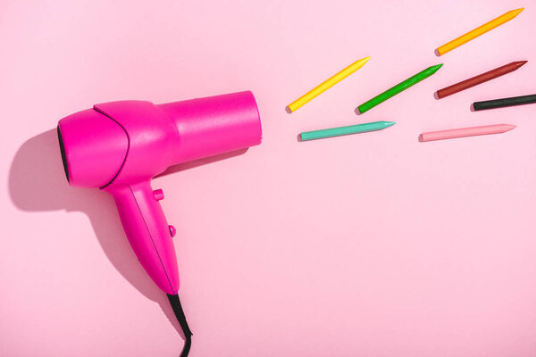 top view of hair dryer blowing colorful crayons on pink