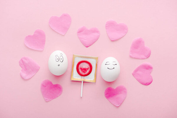 верхний вид яиц со счастливыми и удивленными выражениями лица, бумажными сердцами и презервативами, обернутыми в леденец на розовом
