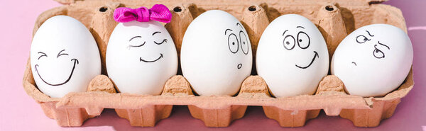 panoramic shot of eggs with different face expressions in egg carton on pink
