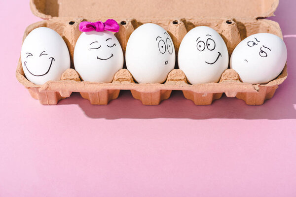 eggs with different face expressions in egg carton on pink with copy space