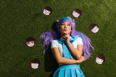 Pensive asian anime girl in purple wig with emoticons lying on grass