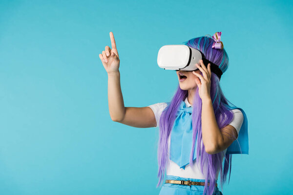 Amazed anime girl in purple wig and vr headset pointing with finger isolated on blue