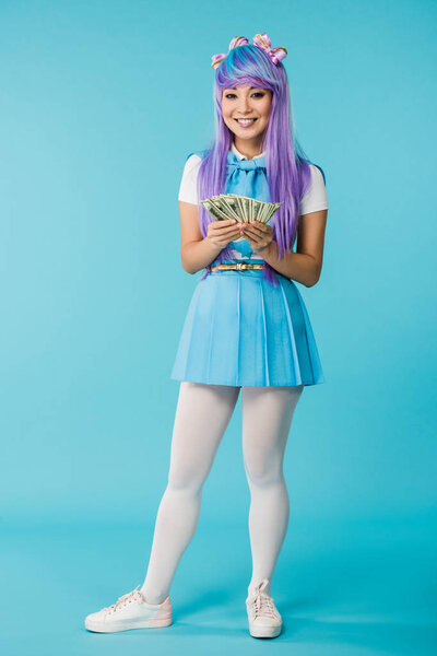 Full length view of asian anime girl in purple wig counting dollar banknotes on blue