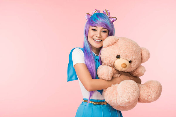 Smiling asian anime girl in wig holding teddy bear isolated on pink