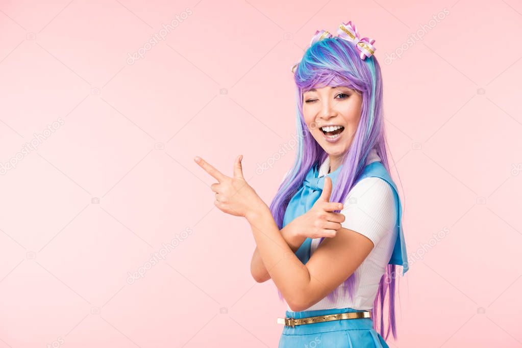 Joyful anime girl in purple wig pointing with fingers and winking isolated on pink