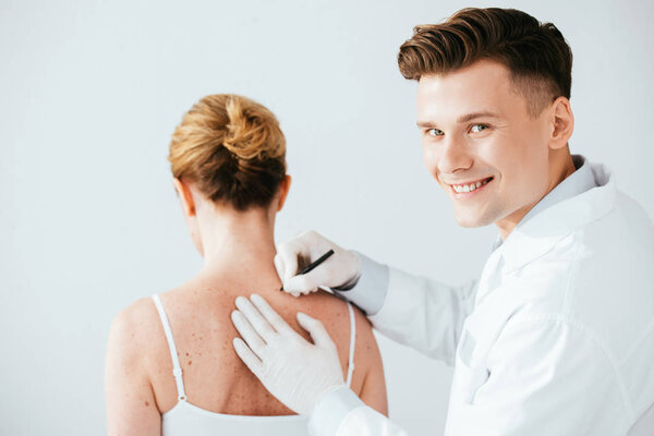 cheerful dermatologist in latex gloves holding marker pen near woman with melanoma isolated on white 