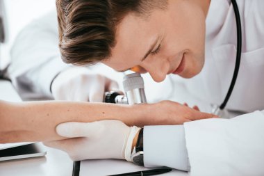 Dermatoskop tutarken kadının elini inceleyen neşeli bir dermatolog. 